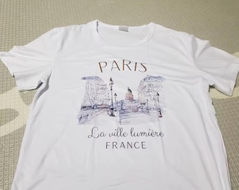 May include: White t-shirt featuring a Parisian street scene illustration in shades of purple, blue, and brown. The word "PARIS" is printed above the illustration, with "La ville lumière FRANCE" below. The shirt has short sleeves and a crew neck.