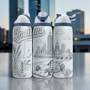 Könnte beinhalten: Drei weiße Wasserflaschen aus Edelstahl mit blauen Deckeln und schwarzen Strichzeichnungen. Die Motive zeigen das Wort Indiana, eine Landkarte, Weizenähren, das Owala-Logo, eine Skyline und einen Indy 500-Rennwagen mit dem Indianapolis Speedway-Logo.