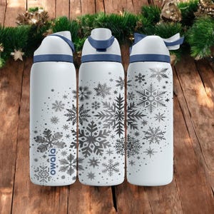 Peut inclure: Trois bouteilles d'eau blanches avec des détails bleus et des motifs de flocons de neige. Les bouteilles ont des couvercles blancs et des poignées bleues. Le mot "owala" est imprimé sur l'une des bouteilles. Les bouteilles sont sur une surface en bois avec des décorations de Noël.