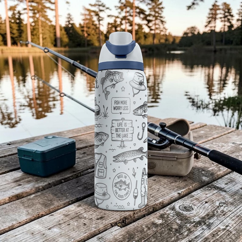 May include: A white water bottle with a blue lid, decorated with black line art of fish, fishing gear, and the phrases "FISH MORE WORRY LESS" and "LIFE IS BETTER AT THE LAKE". The bottle is on a wooden dock near a lake.