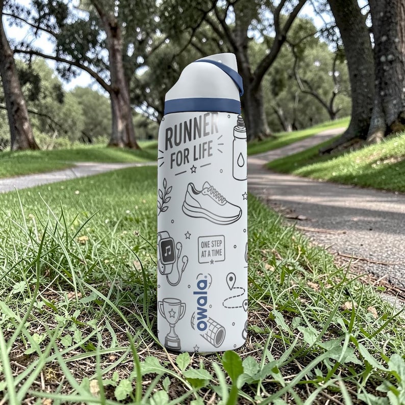 May include: A white and blue Owala water bottle with the text "RUNNER FOR LIFE" and running-themed illustrations. The bottle is standing on a grassy surface with a path and trees in the background.