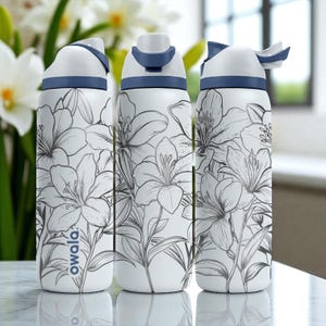 Puede incluir: Tres botellas de agua blancas con un diseño floral. Las botellas tienen una combinación de colores blanco y azul, con un patrón floral en gris. Las botellas tienen una tapa abatible. La marca "owala" está impresa en el lateral.