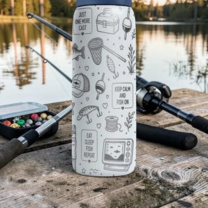 May include: A white insulated water bottle with a navy blue lid, decorated with fishing-themed illustrations and phrases like "Just One More Cast" and "Keep Calm and Fish On." Fishing rods and tackle box are in the background.