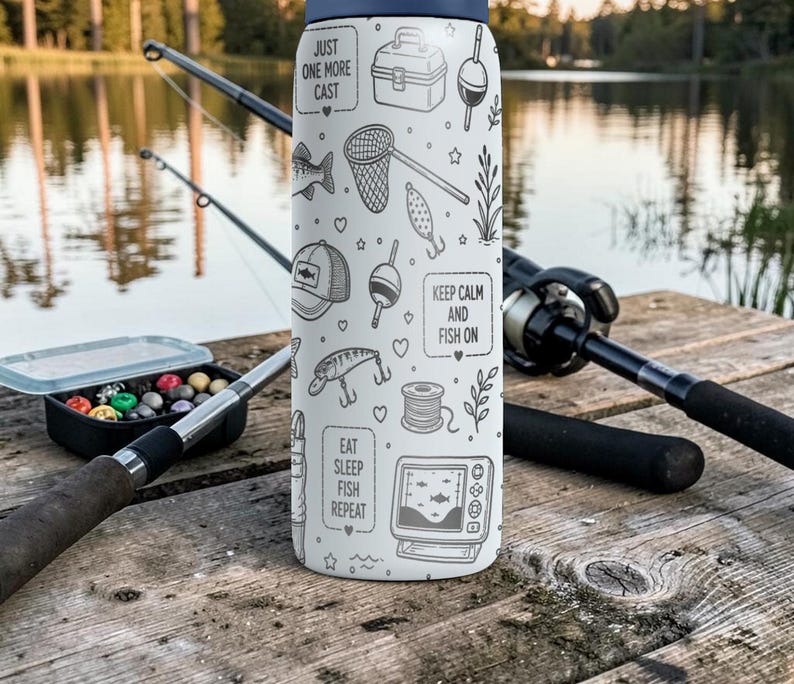 May include: A white insulated water bottle with a navy blue lid, decorated with fishing-themed illustrations and phrases like "Just One More Cast" and "Keep Calm and Fish On." Fishing rods and tackle box are in the background.