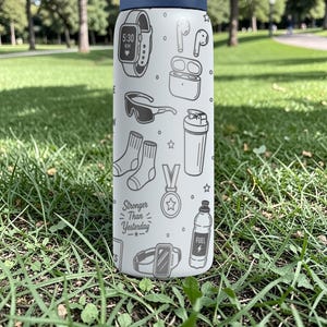 May include: A white water bottle with a navy blue lid, decorated with black and white line drawings of fitness-related items. The bottle features a watch, headphones, socks, and the text "Stronger Than Yesterday."