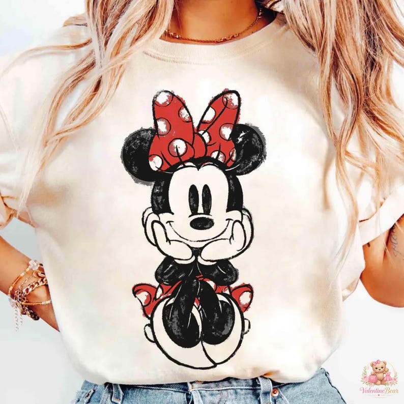 May include: Cream-colored t-shirt featuring a graphic of Minnie Mouse. Minnie is depicted in a seated pose, with a red bow with white polka dots on her head. The design is in black and red, with a vintage, sketched style.