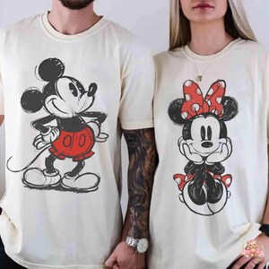 May include: Two cream-colored t-shirts featuring Mickey and Minnie Mouse. Mickey is depicted in a red shorts and Minnie in a red polka dot bow. The cartoon characters are in a sketched style.