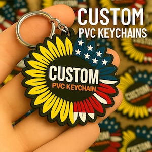 Può includere: Un portachiavi personalizzato in PVC a forma di girasole, tenuto in mano. Il portachiavi presenta petali gialli, stelle blu e bianche e accenti rossi e bianchi. Il testo "CUSTOM PVC KEYCHAINS" è visualizzato in bianco.