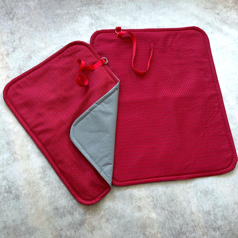 May include: Two rectangular red fabric mats with a subtle white dotted pattern and rounded corners. Each mat features a red elastic loop attached to a metal ring at the top edge. One mat is folded to reveal a smooth, solid gray fabric underside.