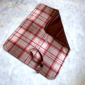 May include: A rectangular mat featuring a beige, white, and red plaid pattern on one side and a solid dark brown fabric on the other. The mat includes a dark brown strap with a metal D-ring attached to the plaid side for securing the item.