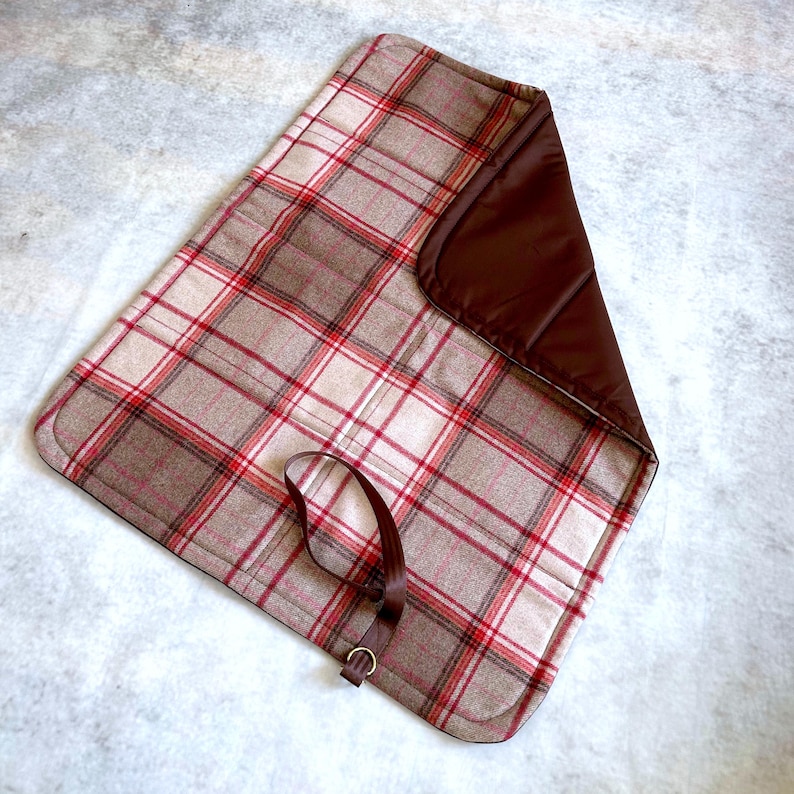 May include: A rectangular mat featuring a beige, white, and red plaid pattern on one side and a solid dark brown fabric on the other. The mat includes a dark brown strap with a metal D-ring attached to the plaid side for securing the item.