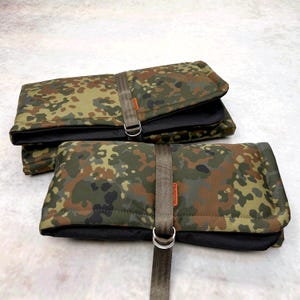 May include: Two rectangular, folded pouches featuring a camouflage pattern in shades of green, brown, and black. Each pouch is secured with a vertical olive-drab nylon strap and a silver-toned metal D-ring, with a small orange brand tag attached.