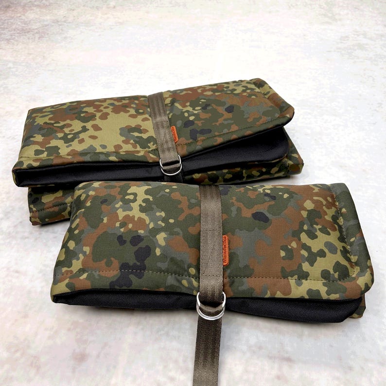 May include: Two rectangular, folded pouches featuring a camouflage pattern in shades of green, brown, and black. Each pouch is secured with a vertical olive-drab nylon strap and a silver-toned metal D-ring, with a small orange brand tag attached.