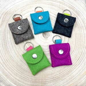 May include: Five small square felt pouches with metal key rings attached to the top. Each pouch features a flap secured by a silver-coloured metal snap button. The pouches are coloured in grey, bright blue, dark charcoal, lime green, and vibrant purple.