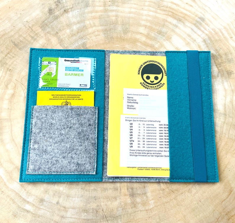 May include: Teal felt folder with a gray felt pocket and a yellow card inside. The card has the text "Kinder-Untersuchungshilfe" and a smiling child's face. The folder has a blue strap.