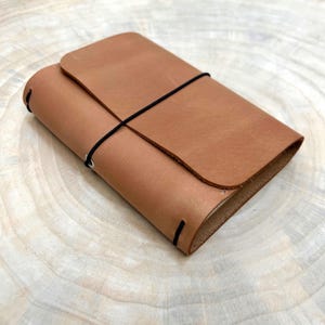 Op de afbeelding: Een notitieboekomslag van bruin leer met een gladde, matte afwerking en een afgeronde flapsluiting. Een dun zwart elastisch koord loopt horizontaal rond het midden van de omslag om de inhoud vast te zetten, terwijl twee kleine zwarte elastische lussen zichtbaar zijn.