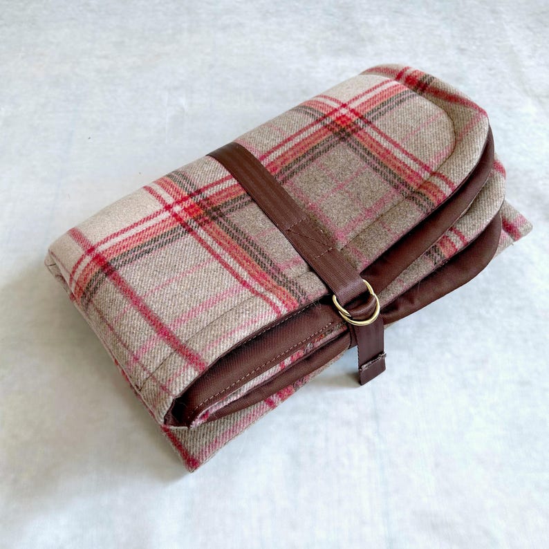 May include: A folded rectangular textile case featuring a beige, red, and grey plaid pattern. The case is secured by a dark brown strap with a gold-toned metal D-ring closure. The interior lining and the strap are made of a solid dark brown material.