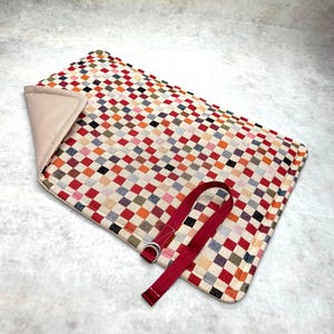 May include: A rectangular mat featuring a multicolored checkered pattern in shades of red, orange, blue, and green on a cream background. The mat has a solid beige lining on one corner and a long, dark red fabric strap with two silver metal rings.