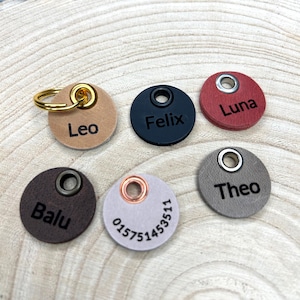 Könnte beinhalten: Fünf runde Tieranhänger aus Leder in Beige, Schwarz, Rot, Dunkelbraun und Hellgrau. Jeder Anhänger hat eine Metallöse und ist mit einem Namen oder einer Nummer graviert: Leo, Felix, Luna, Balu, Theo und 015751453511. Ein goldener Ring ist am beigen Anhänger befestigt.