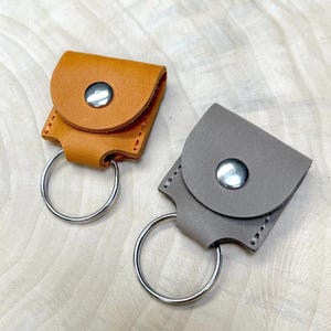 May include: Two small square leather pouches with rounded flaps, one in tan and one in grey. Each pouch features a silver-coloured metal snap button closure and a circular silver-coloured metal split ring attached to the bottom for holding keys.
