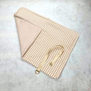 May include: A rectangular fabric mat featuring a beige and white gingham pattern on one side and a solid beige lining on the other. The mat is partially folded to reveal the interior and includes a beige strap with a metal D-ring attached to one edge.