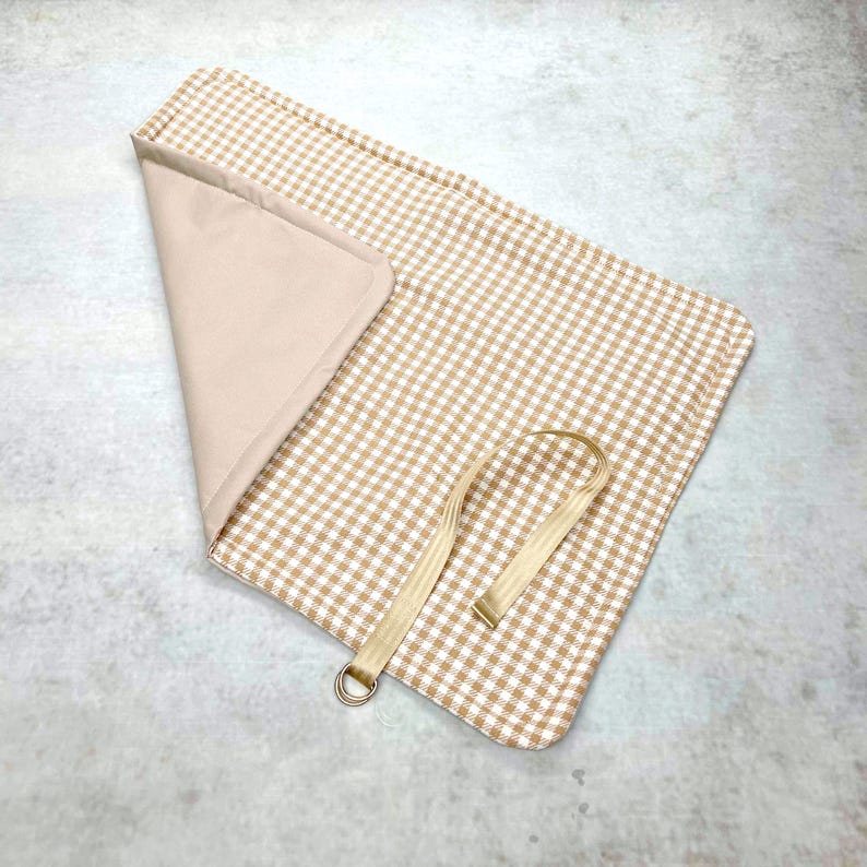 May include: A rectangular fabric mat featuring a beige and white gingham pattern on one side and a solid beige lining on the other. The mat is partially folded to reveal the interior and includes a beige strap with a metal D-ring attached to one edge.