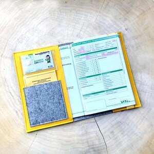 May include: Yellow felt folder with a gray felt pocket and a clear plastic pocket containing a health insurance card. The folder is open to reveal a printed form titled "Fast-Check Geburt (U1) Neugeborenen-Untersuchung".