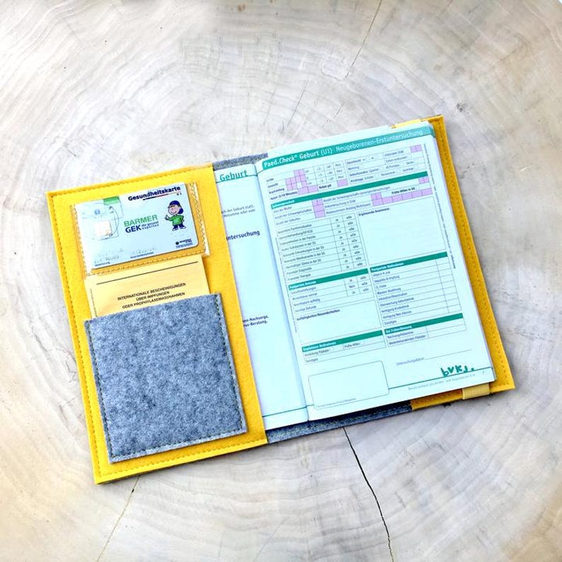 May include: Yellow felt folder with a gray felt pocket and a clear plastic pocket containing a health insurance card. The folder is open to reveal a printed form titled "Fast-Check Geburt (U1) Neugeborenen-Untersuchung".