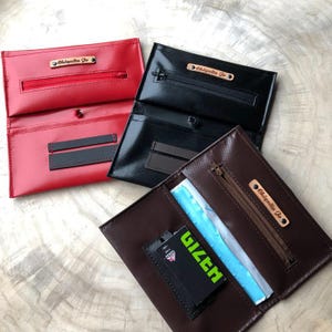 Può includere: Tre bustine per tabacco con etichette personalizzate. Le bustine sono rossa, nera e marrone. La bustina marrone è aperta e contiene un pacchetto di cartine per sigarette.