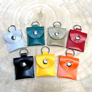 Pode incluir: Um conjunto de sete bolsas quadradas com superfície texturizada, cada uma com um fecho de botão de pressão em metal prateado e um porta-chaves circular em metal preso no topo. As bolsas estão disponíveis em branco, azul-petróleo, bege, vermelho, preto, amarelo e laranja.
