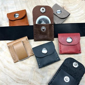 Könnte beinhalten: Sortiment kleiner Lederbeutel in verschiedenen Farben, darunter Braun, Beige, Grau, Schwarz und Rot. Jeder Beutel hat einen Druckknopfverschluss und ist für die Aufbewahrung eines kleinen Gegenstands wie eines Schlüssels oder einer Münze konzipiert.