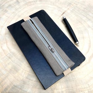 Può includere: Un taccuino nero con un astuccio in tela grigia e una penna nera su una superficie di legno. L'astuccio ha una cerniera argentata e una linguetta beige. La penna ha la punta argentata.