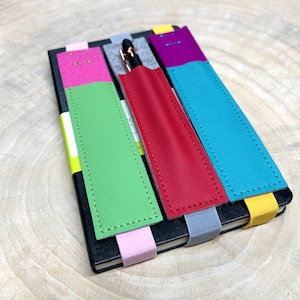 May include: Five colorful leather pen holders attached to a black notebook. The holders are green, red, blue, purple, and pink. Each holder has a matching colored ribbon attached to the bottom.