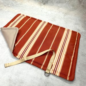 May include: A rectangular mat featuring a rust-orange, white, and tan vertical striped pattern. The mat has a beige underside, a folded corner, and a beige fabric strap attached to a silver-toned metal D-ring for securing the rolled item.