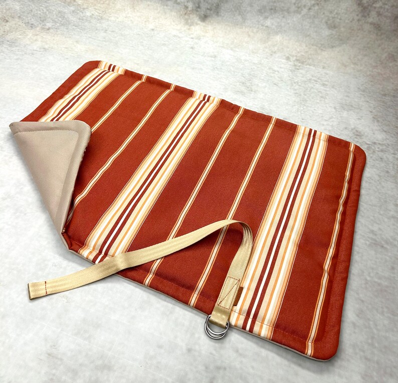 May include: A rectangular mat featuring a rust-orange, white, and tan vertical striped pattern. The mat has a beige underside, a folded corner, and a beige fabric strap attached to a silver-toned metal D-ring for securing the rolled item.