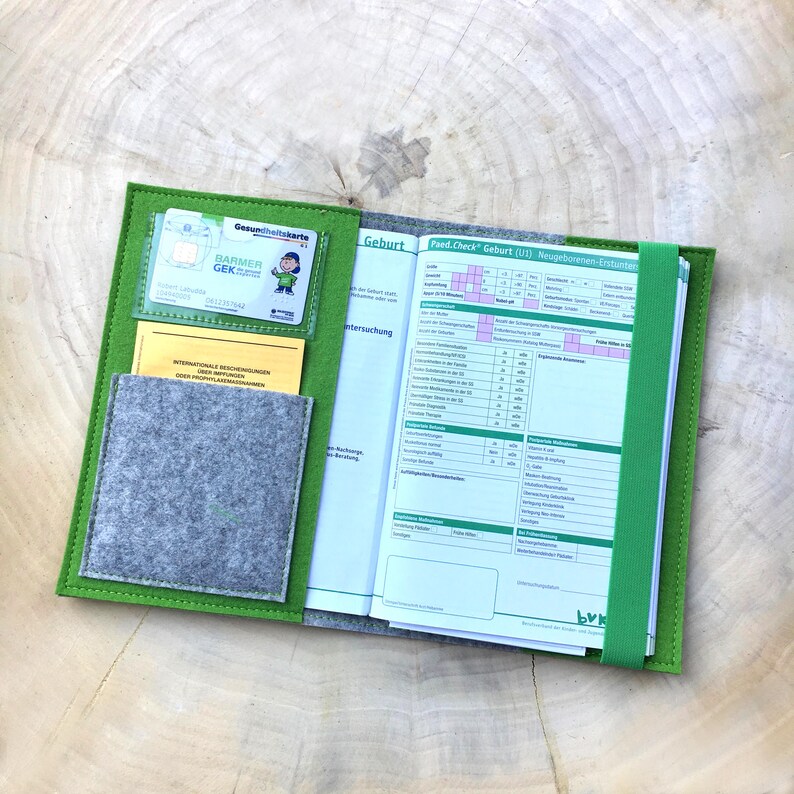 May include: Green felt folder with a clear plastic pocket for a health insurance card, a smaller pocket for a yellow card, and a gray felt pocket. The folder is open to reveal a white page with the title "Geburt" and a chart titled "Fred-Check Geburt (U1) Neugeborenen-Untersuchung".