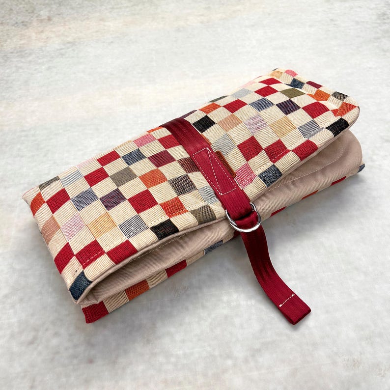 May include: A rectangular fabric organizer case featuring a multicolored checkered pattern in shades of red, blue, yellow, and beige. The case is secured with a dark red fabric strap that includes a silver-toned metal D-ring buckle closure.