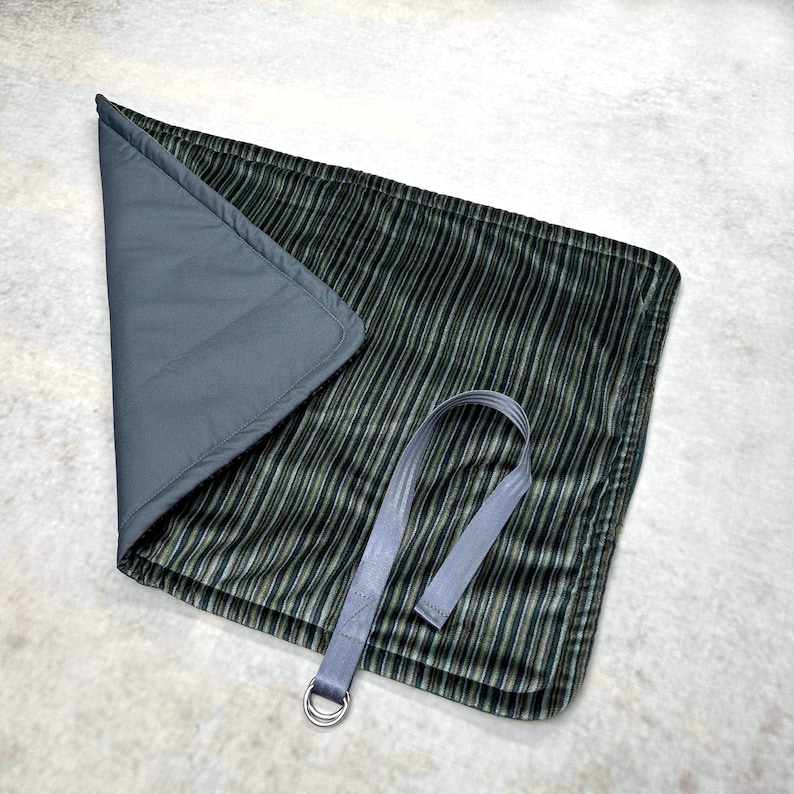 May include: A rectangular fabric mat featuring a dark green and black vertical striped pattern on one side and a solid grey lining on the other. The mat includes a grey fabric strap attached to a silver-toned metal D-ring for secure closure.