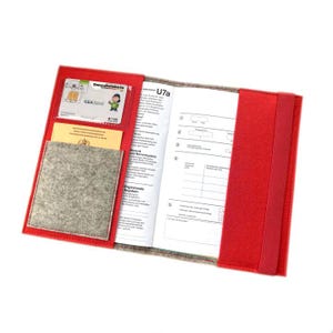 May include: Red felt folder with a gray felt pocket and a clear plastic pocket for a health insurance card. The folder has a label that says "U7a" and includes a section for "Ersprinzende Angaben".