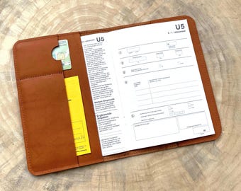 May include: A tan leather document holder featuring a left-side pocket containing a small card and a yellow paper, with an open German U5 medical examination booklet for children aged 6 to 7 months inserted into the right-side sleeve.