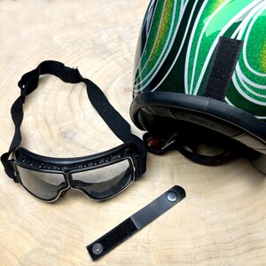 Pode incluir: Óculos de moto de couro preto com lente transparente e um capacete preto com um design de espiral verde e dourado.