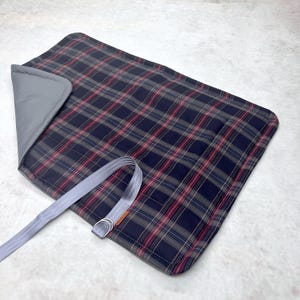 May include: A rectangular quilted mat featuring a dark navy blue, red, and white plaid pattern on one side and a solid grey fabric on the other. The mat includes a grey seatbelt-style strap with a metal D-ring attached for securing the item.
