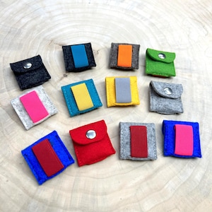 Puede incluir: Una colección de pequeñas bolsas de fieltro coloridas con cierres de botón a presión. Cada bolsa presenta un acento rectangular contrastante en varios colores, incluyendo azul, rosa, naranja y amarillo. Las bolsas están dispuestas sobre una superficie de madera clara.