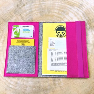 May include: A pink felt folder with a gray felt pocket and a yellow card inside. The card has the text "Kinder-Untersuchung Gemeinsamer Bundesausschuss" and a smiling child icon. The folder has a pink elastic band.