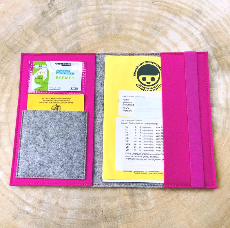 May include: A pink felt folder with a gray felt pocket and a yellow card inside. The card has the text "Kinder-Untersuchung Gemeinsamer Bundesausschuss" and a smiling child icon. The folder has a pink elastic band.