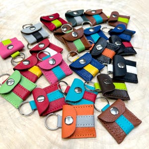 May include: A collection of small, square leather keychains featuring a snap-button closure and a central vertical stripe in a contrasting colour. Each keychain includes a metal split ring and is crafted from textured leather in various colours.