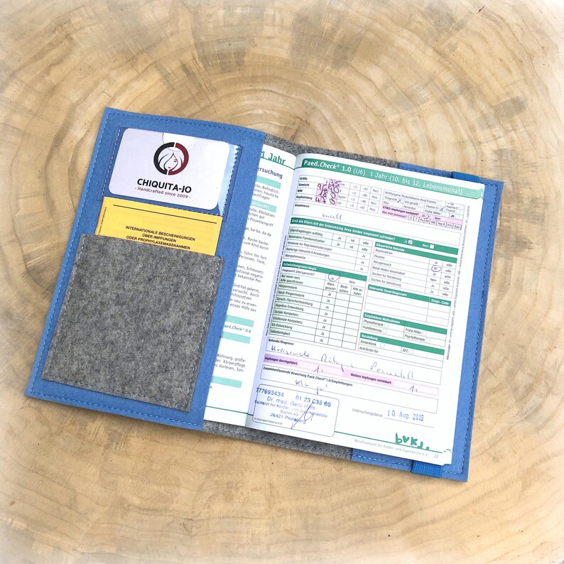 May include: A blue folder with a gray felt pocket and a yellow card with the text "CHIQUITA-JO" in black. The folder is open to reveal a page with a form titled "Paed. Check 1.0 (6)" and the text "1 Jahr" in black.