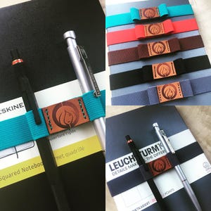 Puede incluir: Dos cuadernos con portaplumas. Los cuadernos son negros y grises con un portaplumas hecho de una banda elástica azul turquesa con una lengüeta de cuero marrón. La lengüeta tiene un diseño estilizado de una figura femenina. El texto en los cuadernos dice "ESKINE" y "LEUCHTTURM19 DETAILS MAKE THE DIFFERENCE".