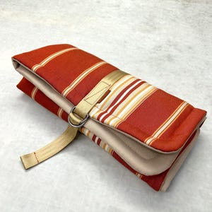 May include: A folded fabric roll featuring a rust-orange and beige striped pattern. The item is secured with a beige nylon webbing strap that feeds through two silver-toned metal D-rings, keeping the folded textile layers firmly in place.