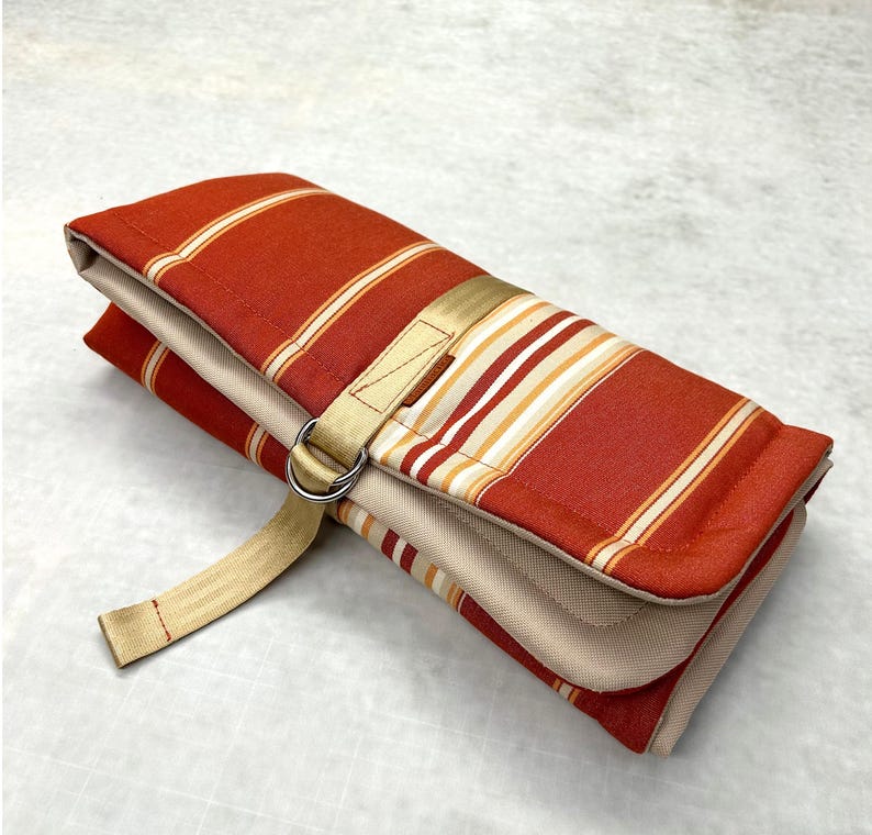 May include: A folded fabric roll featuring a rust-orange and beige striped pattern. The item is secured with a beige nylon webbing strap that feeds through two silver-toned metal D-rings, keeping the folded textile layers firmly in place.